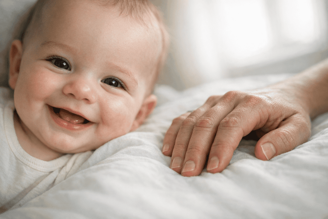 A baby lying on a white bed smiling at the camera while an adult hand rests beside them